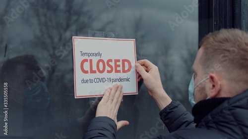 Cuadros en Lienzo Caucasian male wearing medical mask puts a Temporary closed due COVID-19 pandemic sign on a window