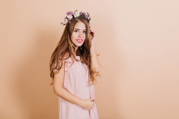 Wall Mural - Amazing lady with long straight hair posing on beige background. European girl with purple flowers on head expressing positive emotions.