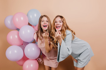 Amazing curly girl in blue dress posing near sister in her birthday. Indoor photo of smiling ladies standing on light background with helium balloons.