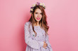 Inspired female model wears purple attire looking to camera. Elegant white girl with beautiful flowers in long hair posing on pink background.