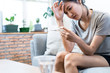 © Kawee - Asian young woman sick with coronavirus. She sit on sofa at home. Girl check temperature with body thermometer herself. Female have fever and headache. People feel sad and lonely. Covid-19 concept