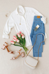 Blue jeans, white shirt, heeled sandals, small bag with chain strap, accessories, pink tulips flowers on beige background. Women's stylish spring summer outfit. Trendy clothes. Flat lay, top view.