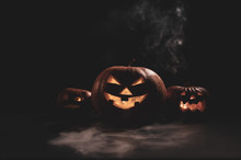 Halloween Card. Jack O Lantern With Candles Glow On A Black Background. A Row Of Creepy Pumpkins With Carved Grimaces Smokes In The Dark.