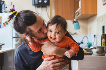 hipster young father hug and kiss his baby infant boy holding on hands. at home on kitchen backgroun