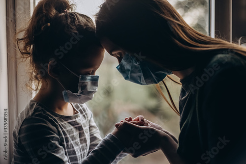 Fotografía Close up of sad young mommy hugs cute preschool daughter both in protective medical masks sits on windowsill and looks out window