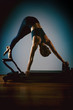 © Georgii - Young girl doing pilates exercises with a reformer bed. Beautiful slim fitness trainer on reformer gray background, low key, art light. Fitness concept