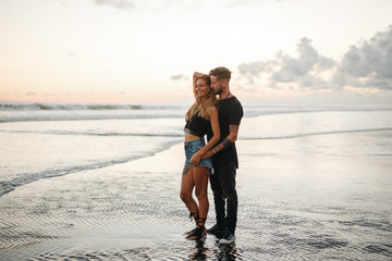 young beautiful guy and the girl posing on the beach and couple in love at sunset gently hug together forever, hipsters, outdoor portrait, close up. Happy couple has fun outdoors on sunset on beach