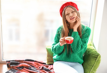 Sticker - Beautiful young woman with cup of coffee sitting on window sill at home