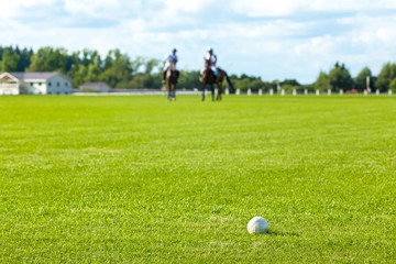 Horse polo field. The ball is in the foreground. Focus on a ball. In the background, two horse polo players in equipment on horseback. They 're out of focus. Forest and cloud sky. Overcast, copyspace