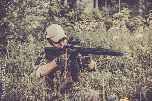 Fotomural A military man or airsoft player in a camouflage suit sits in the grass and aims