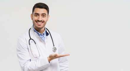 Wall Mural - Horizontal banner of smiling young male doctor showing and presenting something with hand, isolated on gray background with copy space on right side