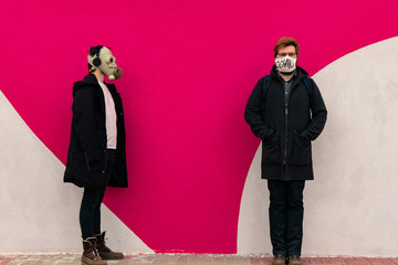 two teenager guy portrait in medicine mask with covid text and gas mask on faces simple posing on concrete painted pink and white wall background texture