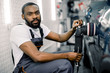 © Sofiia Shunkina - Young handsome bearded African man, worker of auto detailing service, holds a polisher in the hand and polishes the car, looking and posing at camera. Car detailing and polishing concept