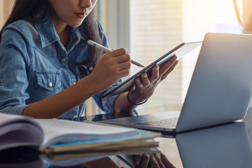 Young asian woman hand holding and using digital tablet, work on laptop computer with book on the desk at home. Online learning, home school concept.