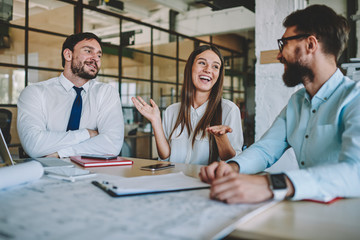 Excited male and female colleagues sitting at table desktop for cooperation brainstorming in modern office interior and laughing during work break for friendly communication, group of happy employees