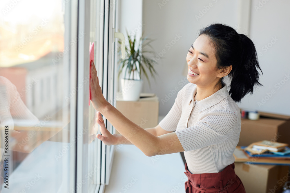 Waist up portrait of young Asian woman washing windows while enjoying Spring cleaning in house or apartment, copy space - obrazy, fototapety, plakaty 
