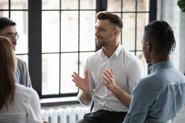 Wall Mural - Happy male psychologist counselling speaking with diverse people sitting in circle at group therapy session. Business coach training staff, having fun, team building activity at work.