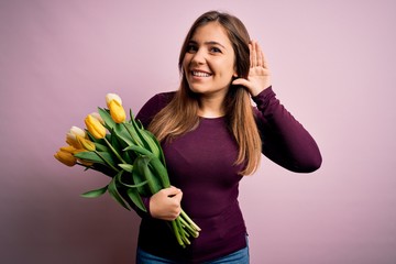 Young blonde woman holding romantic bouquet of yellow tulips flowers over pink background smiling with hand over ear listening an hearing to rumor or gossip. Deafness concept.