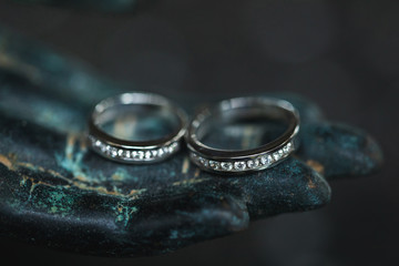 Two white golden wedding rings with diamonds on metal Buddha hand on dark background. Spiritual love concept.