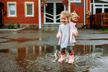 Cute Little Blonde Girl In Pink Jacket, Gray Pants And Rubber Boots Is Jumping Over A Puddle On A Rainy Day