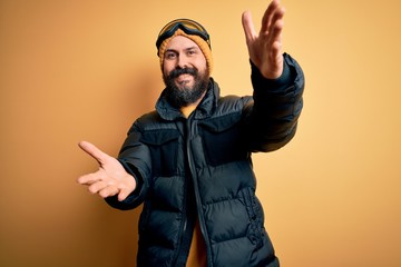 Poster - Handsome skier bald man with beard skiing wearing snow sportswear and ski goggles looking at the camera smiling with open arms for hug. Cheerful expression embracing happiness.