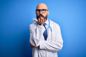 Sticker - Handsome bald doctor man with beard wearing glasses and stethoscope over blue background looking at the camera blowing a kiss with hand on air being lovely and sexy. Love expression.