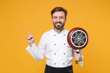 Wall Mural - Smiling young bearded male chef cook or baker man in white uniform shirt posing isolated on yellow background. Cooking food concept. Mock up copy space. Hold frying pan pointing index finger aside up.