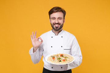 Smiling young bearded male chef cook or baker man in white uniform shirt posing isolated on yellow background. Cooking food concept. Mock up copy space. Hold italian pizza on plate showing OK gesture.