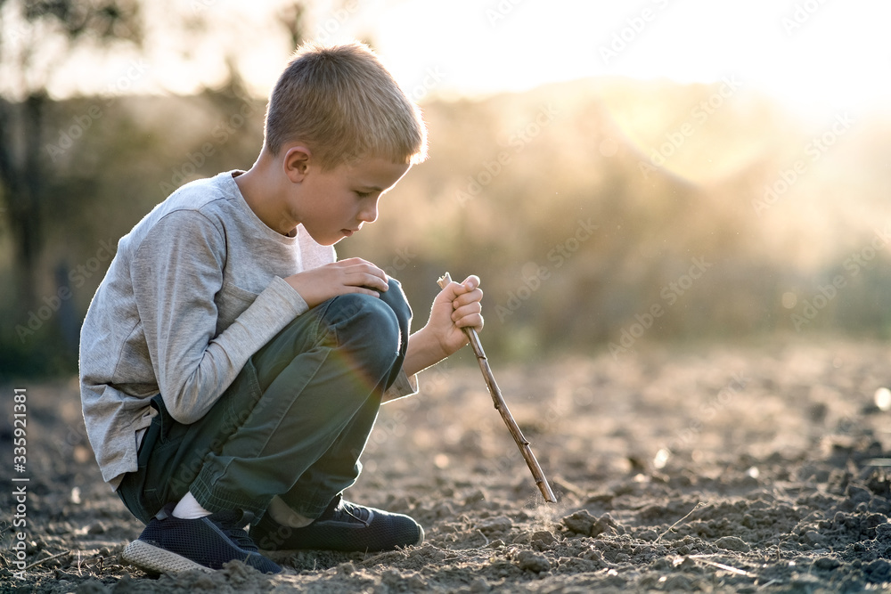 Child boy playing with wooden stick digging in black dirt ground outdoors. - obrazy, fototapety, plakaty 