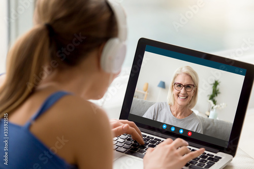 View over shoulder of adult daughter talks by video call with 50s mum. Pc screen view smiling elderly grandmother enjoy virtual chat videoconference with grown up granddaughter. New tech usage concept