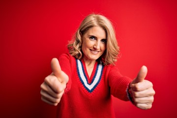 Middle age beautiful blonde woman wearing casual sweater over isolated red background approving doing positive gesture with hand, thumbs up smiling and happy for success. Winner gesture.