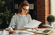 © JenkoAtaman - Young dedicated woman revising papers while working on project at laptop in loft office