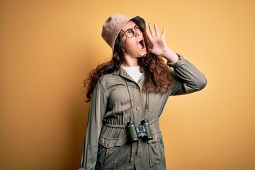 Poster - Young beautiful tourist woman on vacation wearing explorer hat and binoculars shouting and screaming loud to side with hand on mouth. Communication concept.