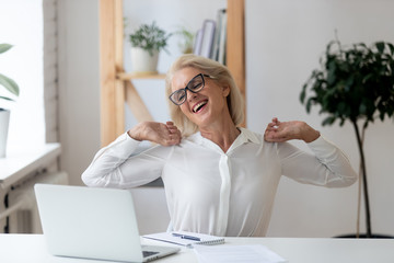 Happy 60 years old businesswoman with hands behind head relaxing in comfortable office chair during break. Smiling female employee with closed eyes resting after work done, leaning back.