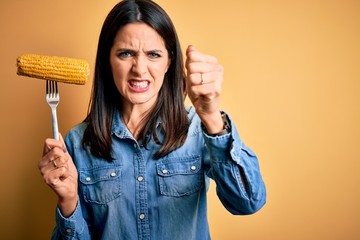 Wall Mural - Young woman with blue eyes holding fork with fresh cob corn standing over yellow background annoyed and frustrated shouting with anger, crazy and yelling with raised hand, anger concept