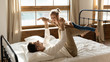 Smiling father lying on comfortable orthopedic mattress bed holding on stretched hands little daughter, practicing funny yoga exercises at home. Happy small girl flying in air, acting as airplane.
