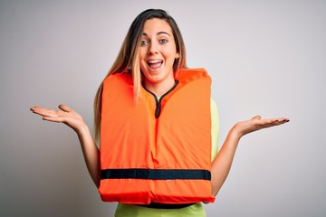 Young beautiful blonde woman with blue eyes wearing orange lifejacket over white background celebrating crazy and amazed for success with arms raised and open eyes screaming excited. Winner concept