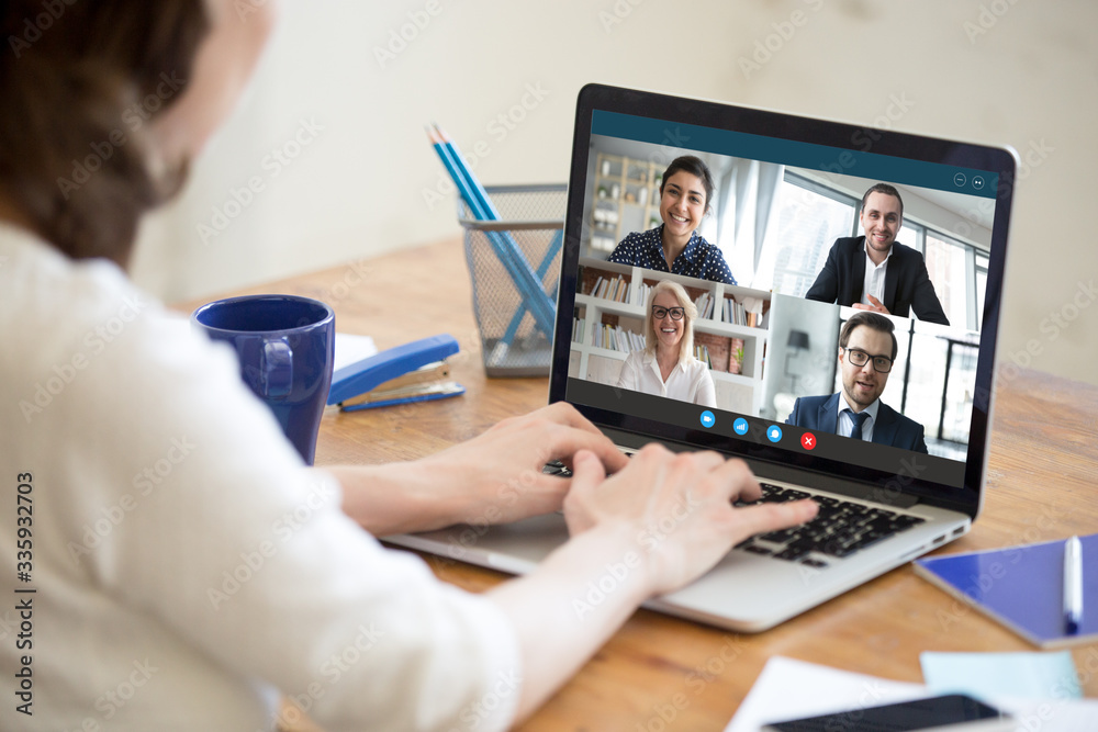 Diverse people take part in group video call pc screen cam app view over woman shoulder, seated at desk. Solve business issues distantly during coronavirus pandemic outbreak, videoconferencing concept - obrazy, fototapety, plakaty 