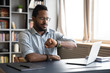 © fizkes - African American young male employee in glasses check time on hand watch waiting for work day to be over, biracial man worker sit at desk use smartwatch at workplace, time management concept