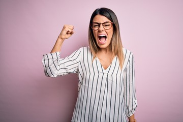 Poster - Young beautiful woman wearing casual striped t-shirt and glasses over pink background angry and mad raising fist frustrated and furious while shouting with anger. Rage and aggressive concept.