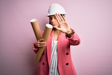 Poster - Young beautiful brunette architect woman wearing safety helmet holding blueprints with open hand doing stop sign with serious and confident expression, defense gesture