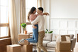 Laughing amazed family couple celebrating real estate purchase. Happy young husband cuddling lifting smiling mixed race wife, having fun among huge cardboard boxed in living room, relocation concept.