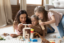 Attractive Young Mother Helping Little Daughters Constructing Building With Wooden Blocks. Smiling Mixed Race Babysitter Playing With Happy Adorable Children Girls On Floor Carpet In Living Room.