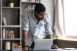 Focused African American male worker in glasses stand at office desk busy working on laptop gadget, concentrated biracial man make notes planning considering business project on computer device