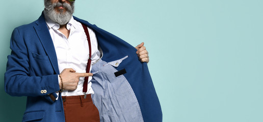 Wall Mural - Cropped photo of man in white shirt, sunglasses, brown pants, suspenders, He pointing at label on the inside of his jacket, posing on blue background