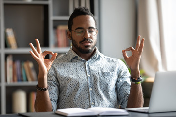 Calm African American man sit at desk meditating relieving negative emotions breathing fresh air, relaxed biracial male distracted from work practice yoga with mudra hands, stress free concept