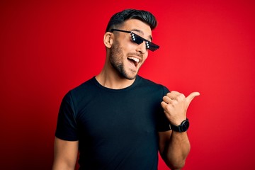 Sticker - Young handsome man wearing funny thug life sunglasses over isolated red background smiling with happy face looking and pointing to the side with thumb up.