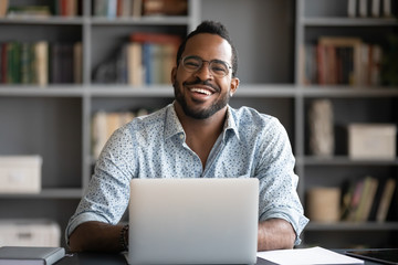 Wall Mural - Portrait of smiling African American man in glasses sit at desk in office working on laptop, happy biracial male worker look at camera posing, busy using modern computer gadget at workplace