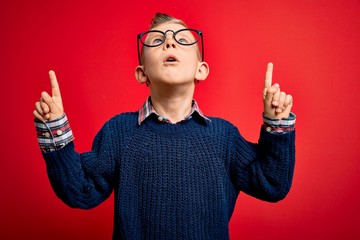 Young little caucasian kid with blue eyes standing wearing smart glasses over red background amazed and surprised looking up and pointing with fingers and raised arms.