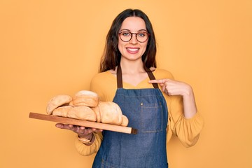 Wall Mural - Young beautiful baker woman with blue eyes wearing apron holding tray with bread with surprise face pointing finger to himself